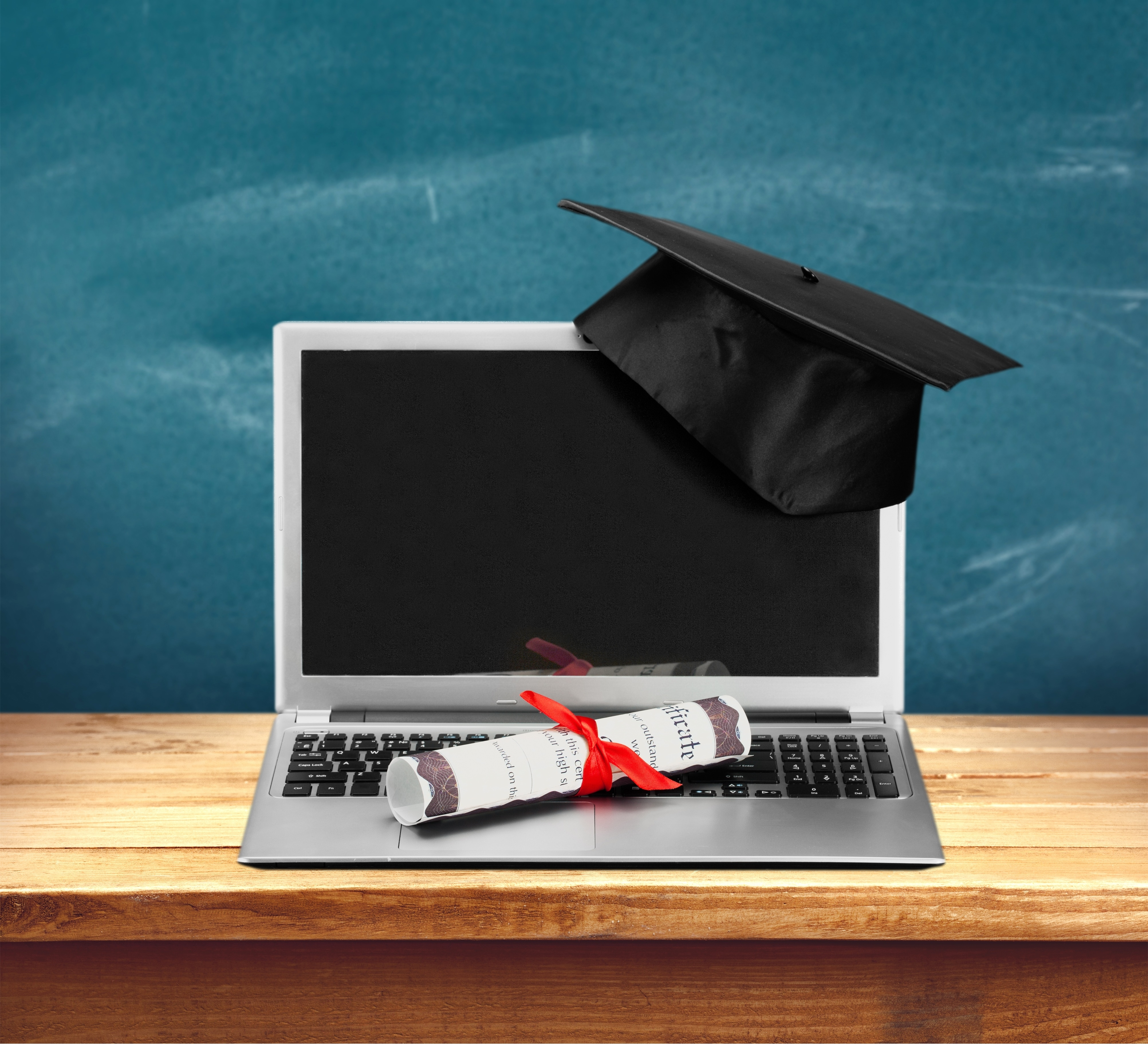 Certificate and graduation cap resting on a computer in front of a chalkboard